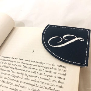 Peut inclure: Un marque-page en cuir noir avec des coutures blanches et une lettre "S" brodée en blanc en écriture cursive.