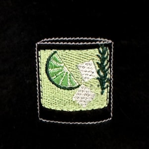Puede incluir: Parche bordado de una rodaja de lima y cubitos de hielo en un vaso, con puntadas verdes y blancas sobre un fondo verde claro.