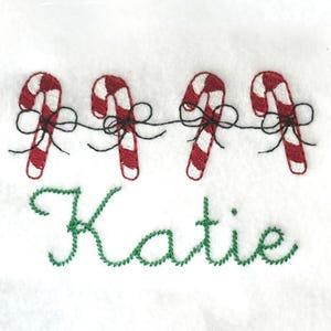 Puede incluir: Diseño bordado con cuatro bastones de caramelo rojos y blancos conectados por hilo negro y lazos. El nombre "Katie" está bordado en verde en cursiva debajo de los bastones de caramelo, sobre un fondo blanco.