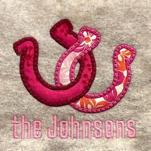 May include: Embroidered design featuring a stylized ampersand in shades of pink and floral patterns, stitched onto a gray fabric. The text "the Johnsons" is embroidered below the ampersand in a matching pink thread, creating a personalized touch.