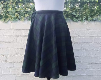 Op de afbeelding: Een zwarte en groene geruite rok met een wikkelontwerp en een zwarte tailleband. De rok is op een mannequin en wordt getoond tegen een witte bakstenen muur.