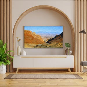 May include: A white media console with three cabinet doors and light wood legs sits beneath a wall-mounted television displaying a landscape painting of orange desert canyons and blue mountains. A green plant and a vase are on the console.