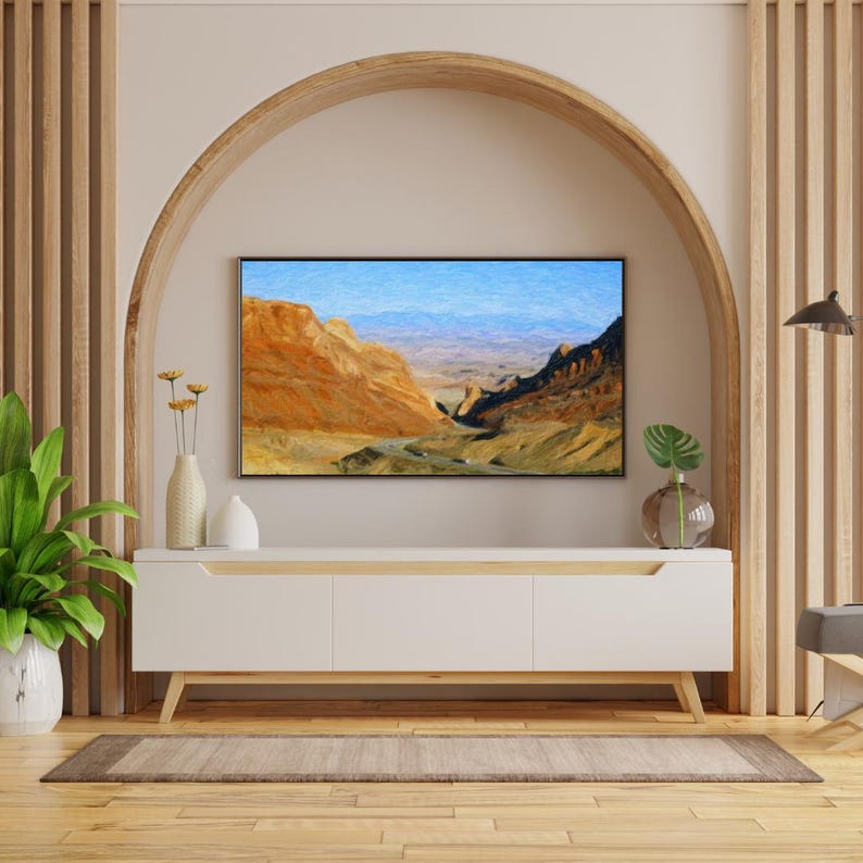 May include: A white media console with three cabinet doors and light wood legs sits beneath a wall-mounted television displaying a landscape painting of orange desert canyons and blue mountains. A green plant and a vase are on the console.