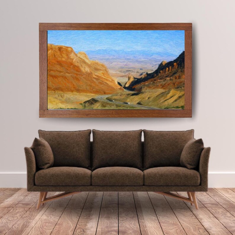 May include: A rectangular landscape painting in a dark wood frame depicting a desert canyon with orange and brown rock formations, a winding road, and distant blue mountains, positioned above a brown three-cushion sofa with two side pillows.