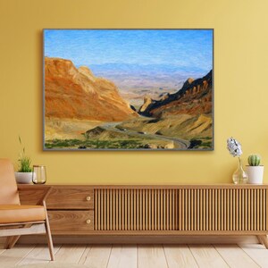May include: A rectangular wall art print featuring a landscape painting of a winding road through a desert canyon with orange and brown rock formations under a bright blue sky. The artwork is displayed in a thin dark frame on a yellow wall.