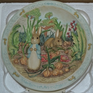 May include: A white ceramic plate with a blue border features a 3D depiction of Peter Rabbit and a second rabbit in a garden setting. The plate is decorated with a detailed design of onions, leaves, and flowers.
