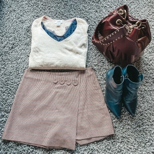 May include: A beige V-neck sweater layered over a dark blue lace camisole, a brown and black houndstooth skort with four decorative buttons, a dark burgundy leather bucket bag with gold studs, and a pair of dark teal pointed-toe ankle boots.