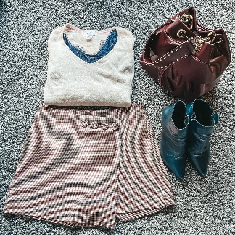 May include: A beige V-neck sweater layered over a dark blue lace camisole, a brown and black houndstooth skort with four decorative buttons, a dark burgundy leather bucket bag with gold studs, and a pair of dark teal pointed-toe ankle boots.