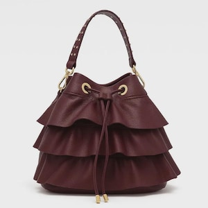 May include: A deep burgundy bucket bag featuring three horizontal ruffled layers of textured leather. The bag includes a drawstring closure with gold-toned metal eyelets and hardware, and a single top handle adorned with small gold-toned metal studs.