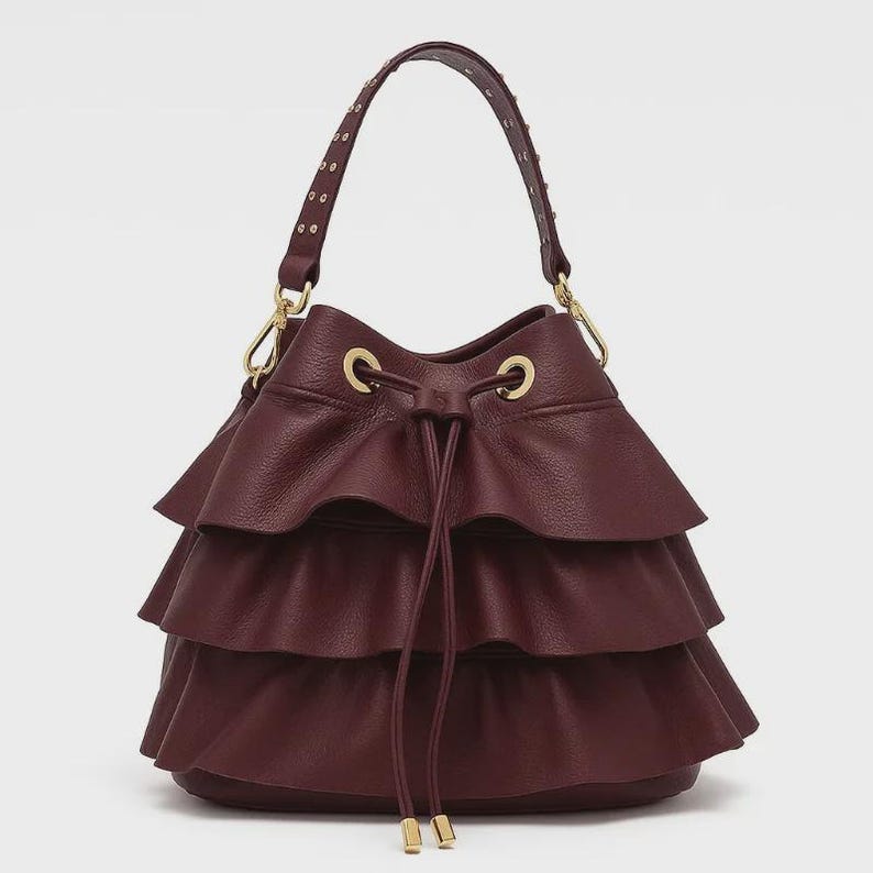 May include: A deep burgundy bucket bag featuring three horizontal ruffled layers of textured leather. The bag includes a drawstring closure with gold-toned metal eyelets and hardware, and a single top handle adorned with small gold-toned metal studs.