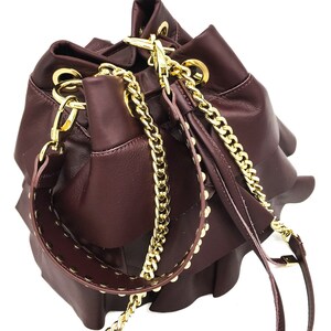 May include: A deep burgundy bucket bag featuring multiple ruffled layers, gold-tone metal hardware, and a detachable shoulder strap. The bag includes a gold chain link strap and a wide leather strap accented with small gold studs along the edges.