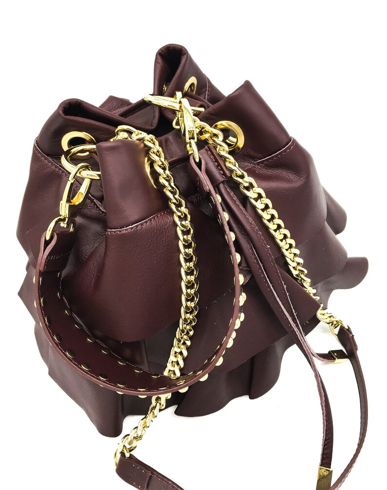 May include: A deep burgundy bucket bag featuring multiple ruffled layers, gold-tone metal hardware, and a detachable shoulder strap. The bag includes a gold chain link strap and a wide leather strap accented with small gold studs along the edges.