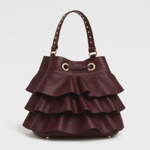 May include: A deep burgundy bucket bag featuring three horizontal ruffled layers of textured leather. The bag includes a single top handle adorned with small gold-tone metal studs and a drawstring closure with gold-tone eyelets at the top opening.