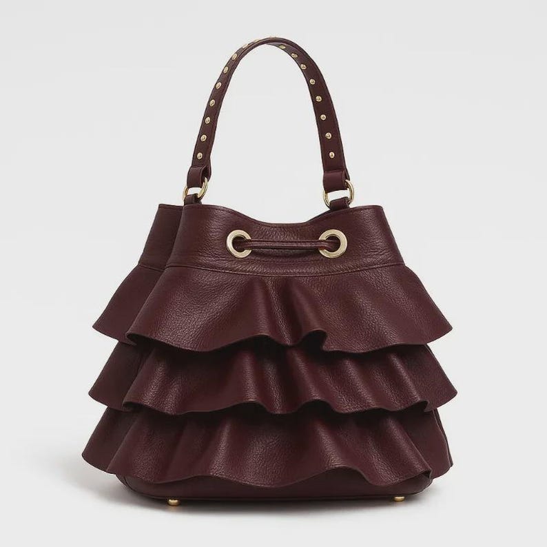 May include: A deep burgundy bucket bag featuring three horizontal ruffled layers of textured leather. The bag includes a single top handle adorned with small gold-tone metal studs and a drawstring closure with gold-tone eyelets at the top opening.