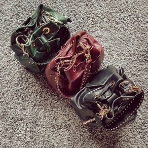 May include: Three bucket bags in dark green, burgundy, and black leather. Each bag features a drawstring closure, gold-tone metal hardware, and a wide shoulder strap embellished with small gold studs along the edges and a gold chain link.
