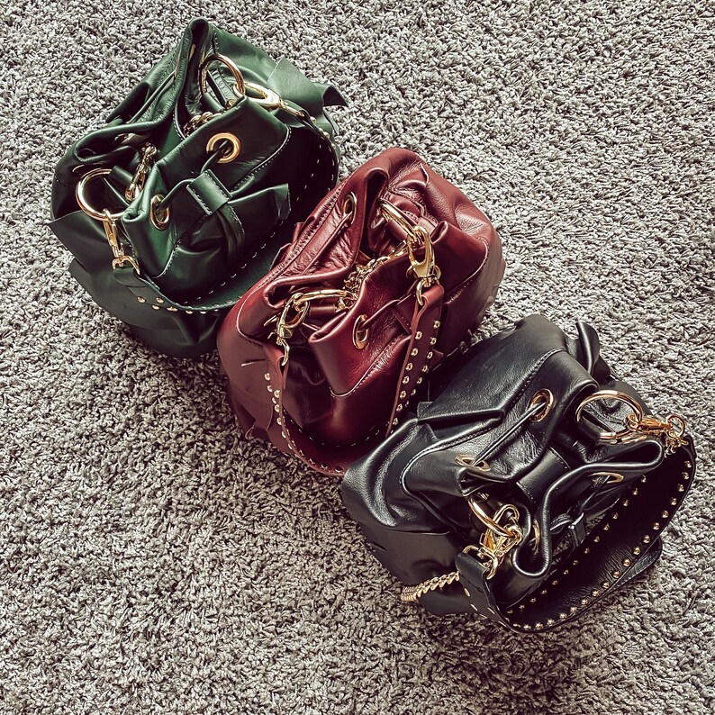 May include: Three bucket bags in dark green, burgundy, and black leather. Each bag features a drawstring closure, gold-tone metal hardware, and a wide shoulder strap embellished with small gold studs along the edges and a gold chain link.