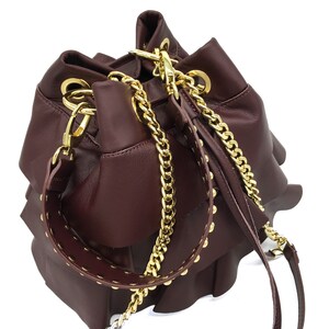May include: A deep burgundy leather bucket bag featuring tiered ruffled layers, a gold-tone chain shoulder strap, and a matching leather strap adorned with gold-tone metal studs. The bag includes gold-tone hardware rings and lobster clasps.
