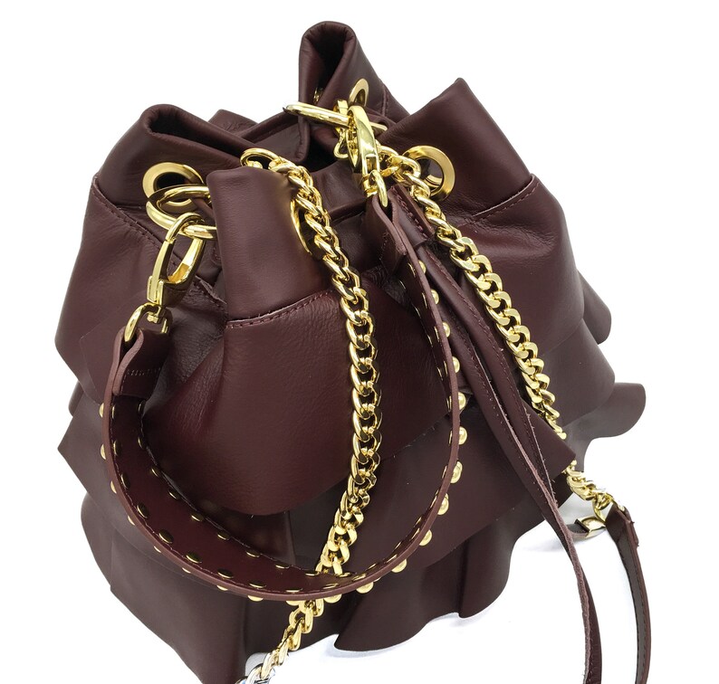 May include: A deep burgundy leather bucket bag featuring tiered ruffled layers, a gold-tone chain shoulder strap, and a matching leather strap adorned with gold-tone metal studs. The bag includes gold-tone hardware rings and lobster clasps.