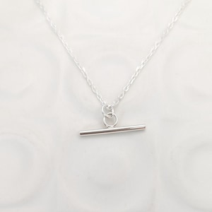 May include: A silver-toned necklace featuring a delicate chain and a horizontal bar pendant attached to a small circular jump ring. The metal has a polished, reflective finish, and the design is simple, consisting of a thin chain and a straight bar.