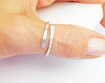 Puede incluir: Un anillo de pulgar plateado con un diseño minimalista. El anillo se envuelve alrededor del pulgar con dos bandas paralelas ligeramente aplanadas. El metal tiene una textura cepillada. El anillo se muestra en el pulgar de una persona.