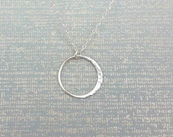 May include: A silver-colored necklace featuring a delicate chain and a circular pendant. The pendant has a smooth finish on one side and a textured, hammered surface on the other, creating a subtle contrast in light reflection across the metal.