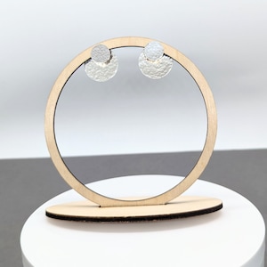 May include: Silver-coloured stud earrings with a hammered texture, displayed on a wooden jewellery stand. The earrings have a small, round stud above a larger, semi-circular shape. The stand is light brown in colour.