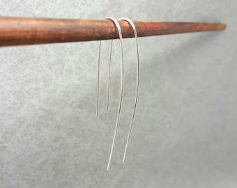 May include: A pair of long, thin, silver-colored U-shaped earrings hanging from a horizontal wooden rod. The earrings feature a smooth, curved wire design with two parallel ends of varying lengths, creating a simple and elongated silhouette.