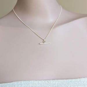 Op de afbeelding: Een delicate zilveren ketting met een kleine staafhanger. De ketting wordt getoond op een mannequin, waarbij het minimalistische ontwerp wordt benadrukt. De ketting is fijn en de staaf is horizontaal, wat een eenvoudige, elegante uitstraling creëert.