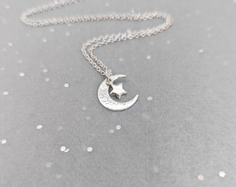 May include: A silver necklace with a crescent moon charm and a small star charm. The necklace is on a gray background with silver glitter.