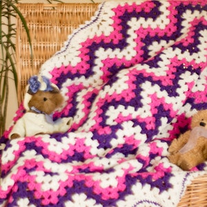 May include: A crocheted blanket with a chevron pattern in white, pink, and purple. Two teddy bears are sitting on the blanket.