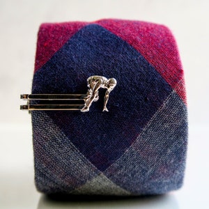 May include: A silver tie clip featuring a stylized diver in a diving pose. The tie clip is on a red and blue plaid tie.