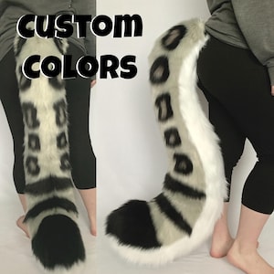 May include: A faux fur tail in a spotted pattern with black, white, and gray colors. The tail has a rounded tip and is shown against a white background. The text "CUSTOM COLORS" is displayed in black and white.