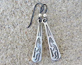 May include: A pair of silver dangle earrings featuring a Native American design. The earrings are suspended from black hooks.