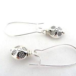 May include: A pair of silver earrings featuring a floral design. The earrings are made from silver wire and have a small, round, silver charm with a floral pattern.