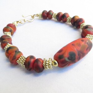 May include: A bracelet with red, orange, and black marbled beads separated by gold beads. The bracelet has a gold clasp.