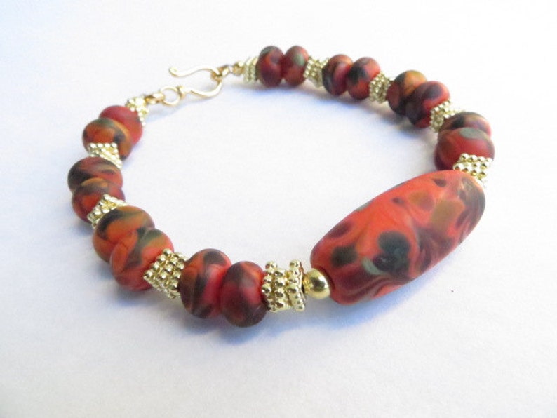 May include: A bracelet with red, orange, and black marbled beads separated by gold beads. The bracelet has a gold clasp.