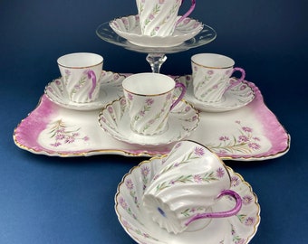 May include: A vintage teacup set featuring white porcelain cups and saucers with a spiral design, purple handles, and gold trim. The set includes a serving tray with a pink floral border and a glass cake stand. The teacups and saucers are decorated with purple flowers.