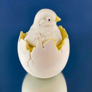 May include: A white ceramic chick figurine inside a cracked white egg with yellow accents. The chick is looking to the right.