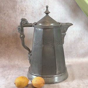 May include: A silver pitcher with an ornate design and a lid. The pitcher has a handle with a decorative scroll design. Two lemons are in the foreground.
