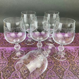 Pode incluir: Seis copos de vinho de vidro transparente com desenhos gravados em um pano de mesa estampado em roxo e dourado. Os copos têm uma base redonda e uma taça larga.