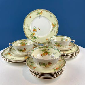 Puede incluir: Un juego vintage de boles de sopa y platillos con un patrón floral. Los boles tienen dos asas y los platillos tienen un borde festoneado. El patrón presenta un fondo verde claro con flores naranjas, amarillas y azules.