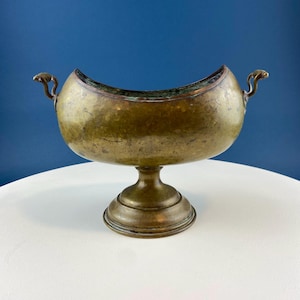 May include: A vintage brass bowl with a half-moon shape and two handles. The bowl sits on a pedestal base.