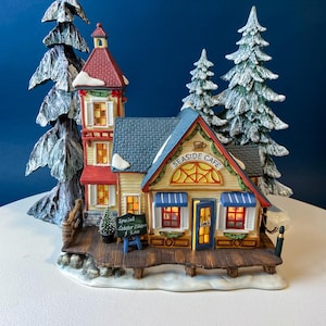 May include: A miniature Seaside Cafe building with a tall red-roofed tower, a blue-roofed main structure, and a wooden deck. The cafe features illuminated windows, a sign reading Special Lobster Dinner $3.00, and two snow-dusted evergreen trees.