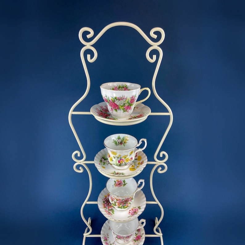 May include: White metal tiered stand with four floral teacups and saucers. The stand has a decorative scrolled design.
