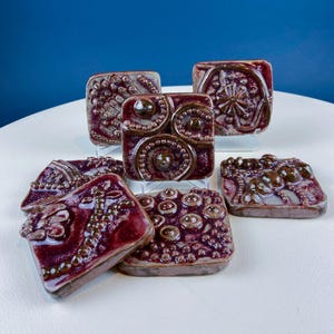 May include: Six ceramic tiles with a burgundy glaze and raised patterns. The patterns include circles, dots, and swirls. The tiles are arranged in a staggered pattern on a white surface.