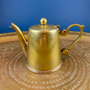 May include: A gold-colored brass teapot with a lid and a handle. The teapot is sitting on a decorative tray with a gold-colored background.