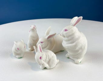 May include: Five white porcelain bunny figurines with pink eyes. The bunnies are in various poses, including sitting, standing, and lying down.