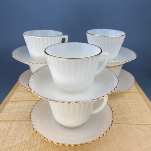 May include: A stack of four white milk glass teacups and saucers with gold trim. The cups and saucers have a fluted design.