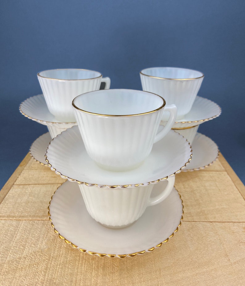 May include: A stack of four white milk glass teacups and saucers with gold trim. The cups and saucers have a fluted design.