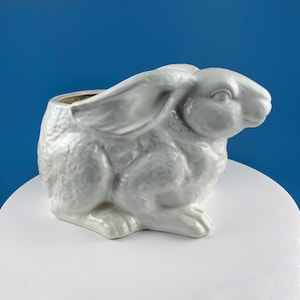 May include: A white ceramic rabbit planter with a hollowed-out interior. The bunny is in a seated position with its ears perked up. The planter is on a white surface with a blue background.